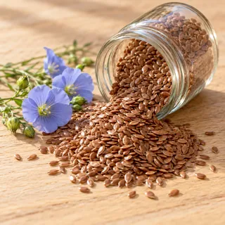 Brown flaxseeds spilling from a glass jar onto a wooden table, with blue flax flowers